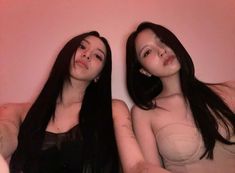 two women with long black hair are sitting on a pink couch and posing for the camera