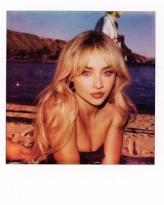 a woman with long blonde hair sitting on the beach next to water and looking at the camera