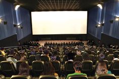 an auditorium full of people sitting in chairs watching a movie on the big screen at the end of the hall