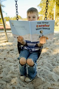 a young boy reading a book on a swing in the sand with chains hanging from it's sides