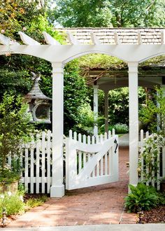 a white fence and gate in the middle of a brick walkway surrounded by greenery