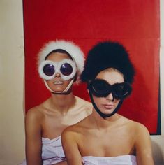 two women in white dresses and black sunglasses are sitting on a bed with red wall behind them