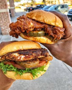 two hands holding sandwiches with meat and veggies on them in front of a parking lot