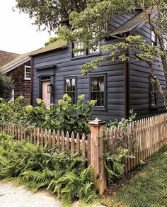 a black house with a picket fence and trees in the front yard, surrounded by greenery