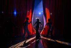 some people are dancing on stage in front of red curtained curtains with lights coming from behind them