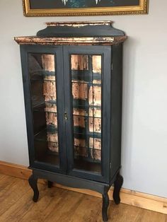 an old black cabinet with glass doors and wooden legs in front of a painting on the wall