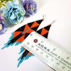 a pair of orange and black beaded earrings next to a ruler with flowers in the background