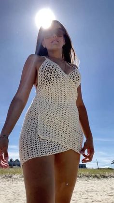 a woman in a white crochet dress walking on the beach with her hands behind her back