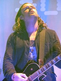 a man with long hair holding a guitar and looking up at the sky in front of him