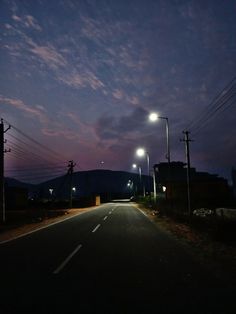 an empty road with street lights and telephone poles in the distance at night, under a cloudy sky