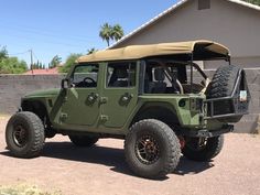 a green jeep is parked in front of a building with a tan roof and two large tires