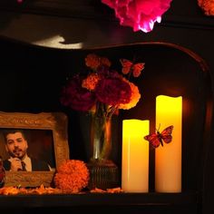 flowers and candles are on a mantle with a framed photo in the middle, next to two lit candles