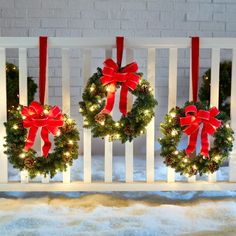 three christmas wreaths with red bows hanging from them on a white rail in front of a brick wall