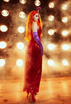 a woman in a red dress and purple gloves standing on a wooden floor with lights behind her