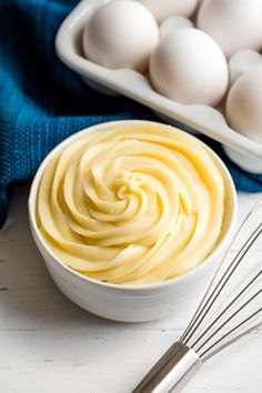 a bowl filled with mayonnaise next to an egg whisk on a white table