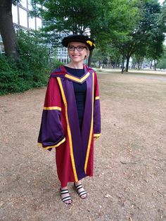 a woman wearing a purple and yellow graduation gown standing in the grass with trees behind her