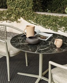 an outdoor table with two chairs and a coffee cup on it, next to a potted plant