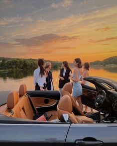 four women standing on the back of a convertible car in front of a lake at sunset