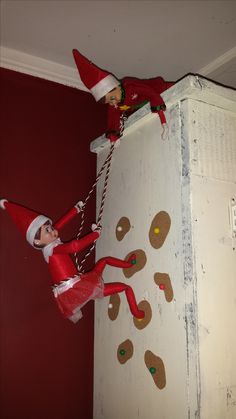 two elfs on top of a refrigerator with christmas decorations hanging from the door and one is wearing a santa hat