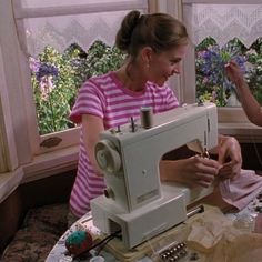 two women are working on sewing machines in a room with large windows and purple flowers