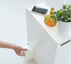 a person holding a roll of toilet paper in front of a white cabinet with a potted plant on top