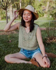 a woman sitting in the grass wearing a hat and holding her hand on her head