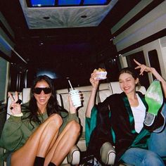 two women sitting in the back of a limo eating and drinking beverages while holding up peace signs