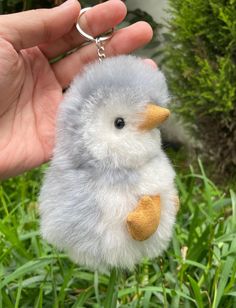 a hand holding a tiny gray and white chick keychain in the grass with it's foot