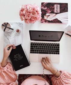 a woman sitting at a desk working on her laptop computer with pink flowers and books nearby