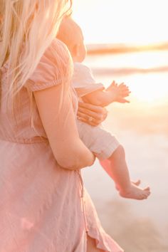 a woman holding a baby in her arms on the beach at sunset or sunrise time