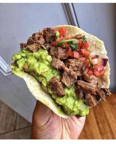 a hand holding a taco with meat, guacamole and tomatoes on it