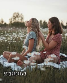 two women sitting on a blanket in the middle of a field with daisies around them