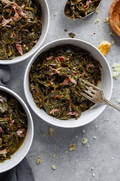 three white bowls filled with cooked greens on top of a gray surface next to two silver spoons