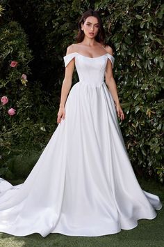 a woman in a white wedding dress standing next to some bushes and flowers with her hands on her hips