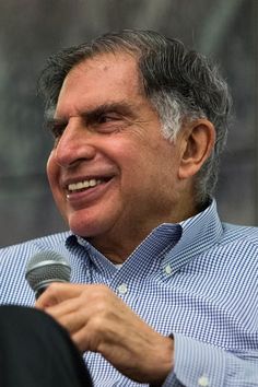 a smiling man holding a microphone in his right hand and wearing a blue checkered shirt