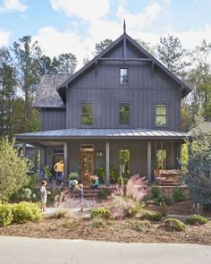 two people are standing in front of a gray house with lots of trees and bushes