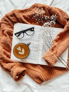 an open book, coffee cup and glasses on top of a white bed with orange blanket