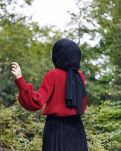 a woman wearing a red sweater and black pleated skirt is standing in the woods