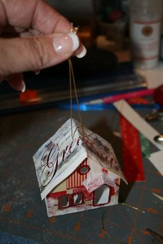 a person holding a small house ornament on a table with scissors and other crafting supplies