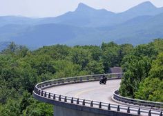 two people riding motorcycles down a winding road in front of trees and mountain range with mountains in the background