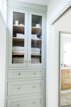 a white cabinet with glass doors and drawers in a bathroom next to a large mirror