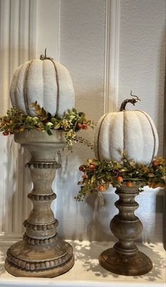 two white pumpkins are sitting on top of some vases with leaves and berries