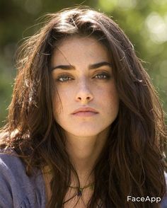 a woman with long brown hair and green eyes looks into the distance while standing in front of trees