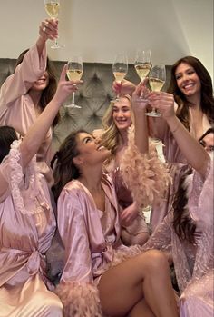 four women in pink robes toasting with wine glasses while sitting on a bed together