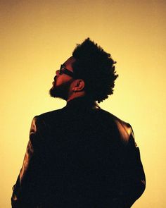 a man with curly hair and glasses standing in front of a yellow background wearing a black jacket