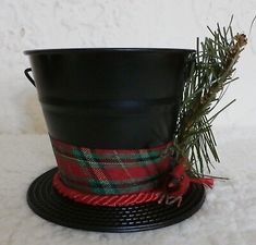 a black pot with a red and green plaid ribbon on the rim next to a pine branch