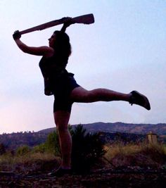 a woman holding a baseball bat while standing on top of a dirt field next to a forest