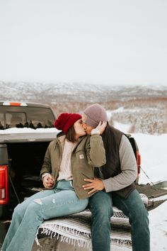 a man and woman sitting on the back of a pickup truck in the snow kissing