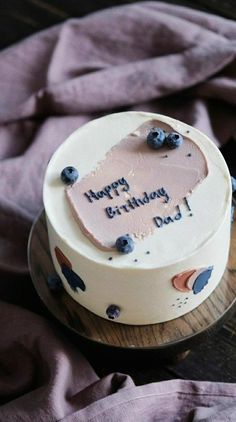 a birthday cake with frosting and blueberries is on a wooden table next to a purple blanket