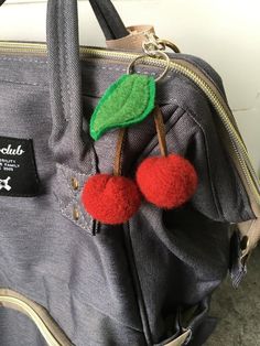 two cherries are attached to the handle of a bag that is gray and has a red pom - pom on it